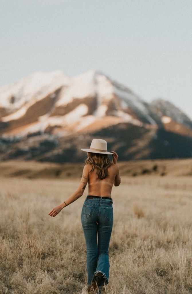 a woman in jeans and a hat walking through a field with mountains in the background