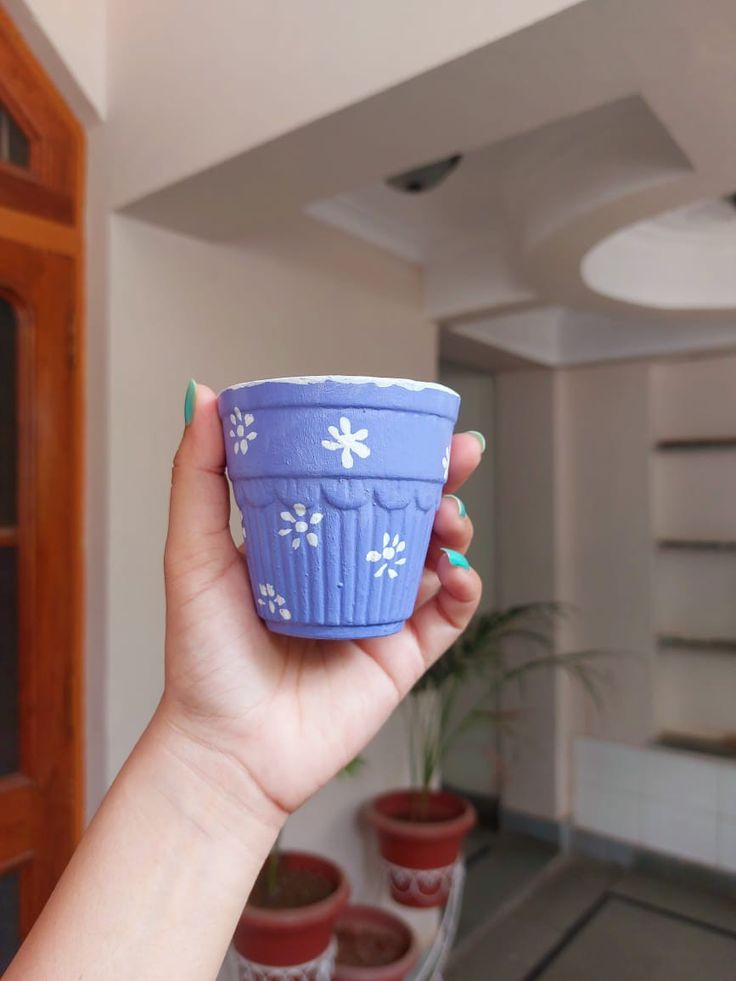 a hand holding a small blue cup with white flowers on it in front of some potted plants