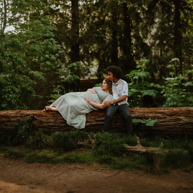 a man and woman are sitting on a log in the woods, hugging each other