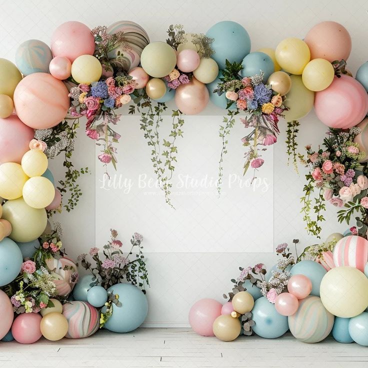 an arrangement of balloons, flowers and greenery in front of a white backdrop for a baby shower