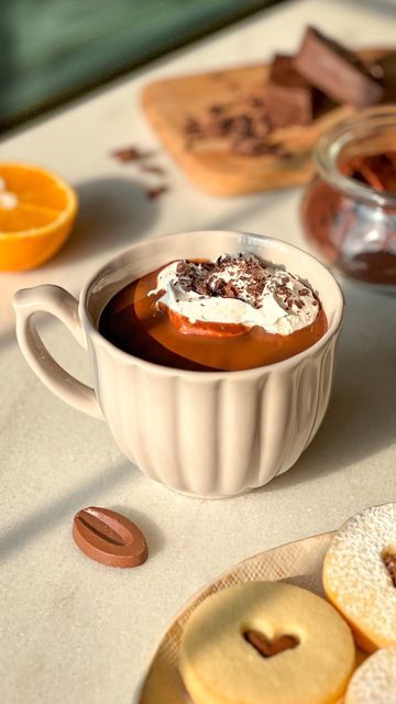 a cup of hot chocolate with whipped cream and orange slices on the table next to it