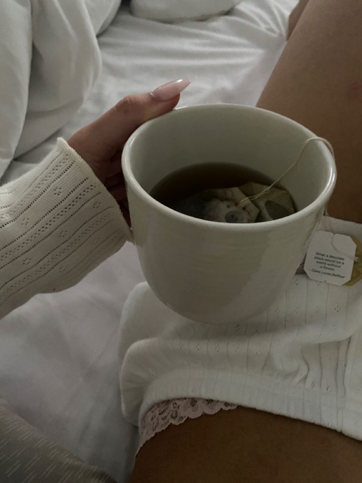 a woman is holding a cup of tea in her hand while sitting on the bed