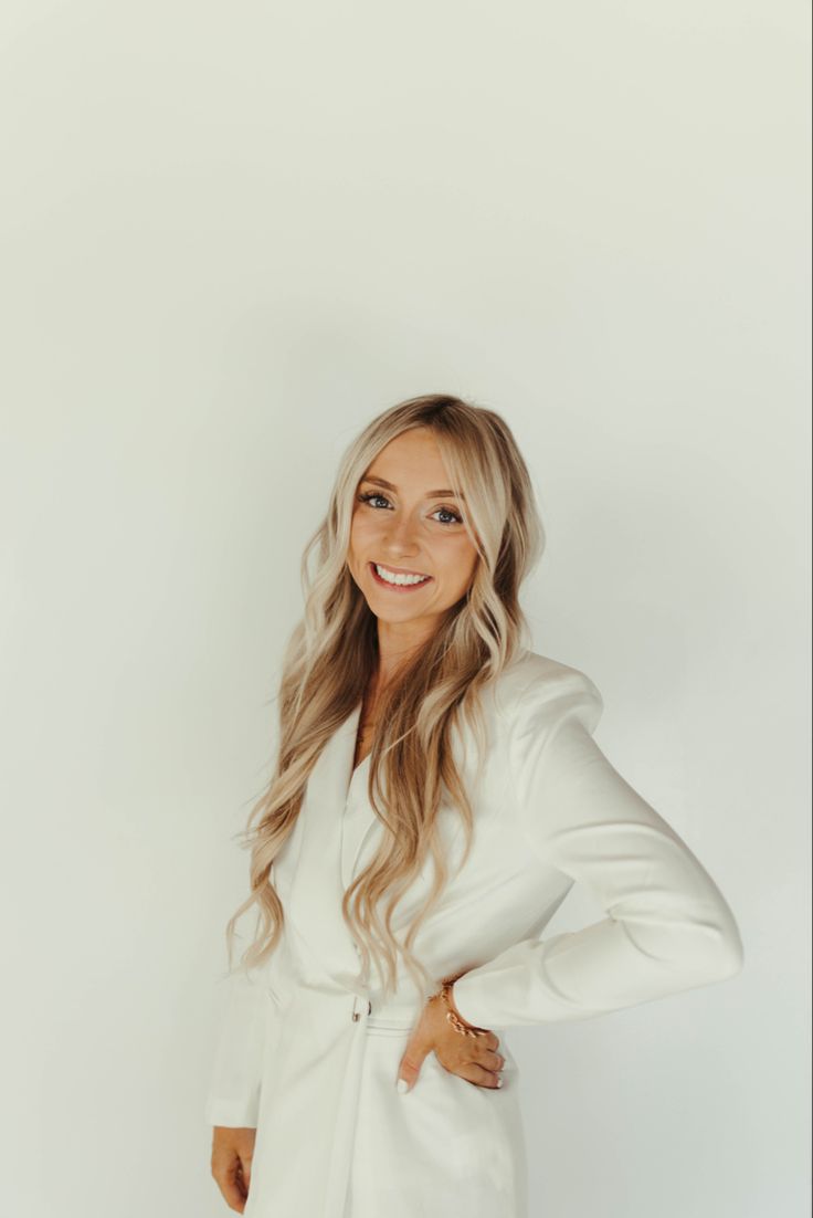 a woman with long blonde hair wearing a white blazer and jeans, smiling at the camera