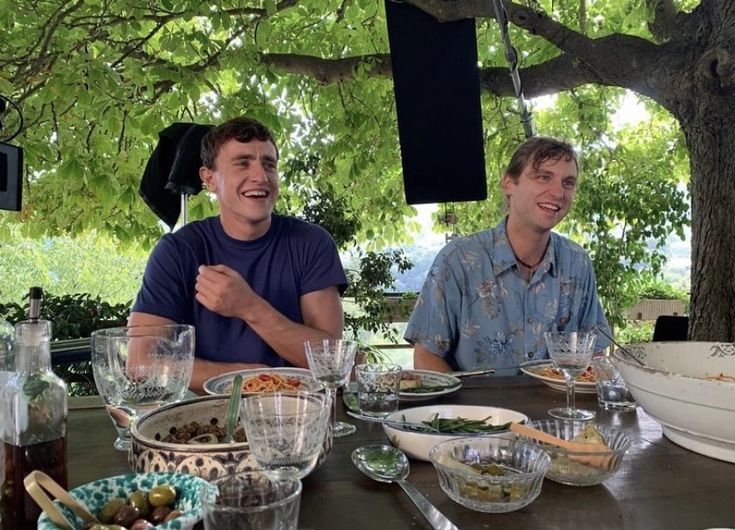 two men sitting at a table with food and drinks in front of them, under a tree