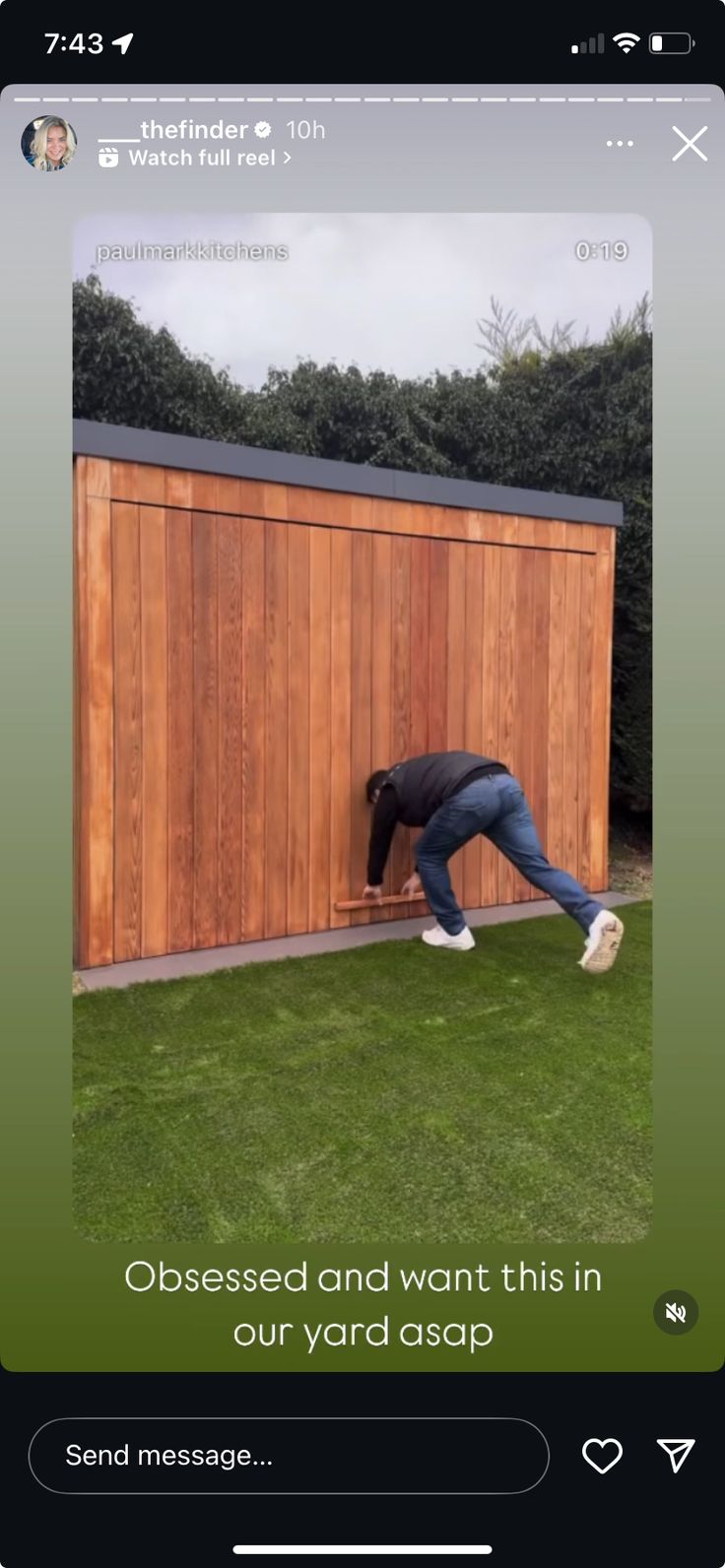 a man leaning over to pick up something from the grass in front of a wooden shed