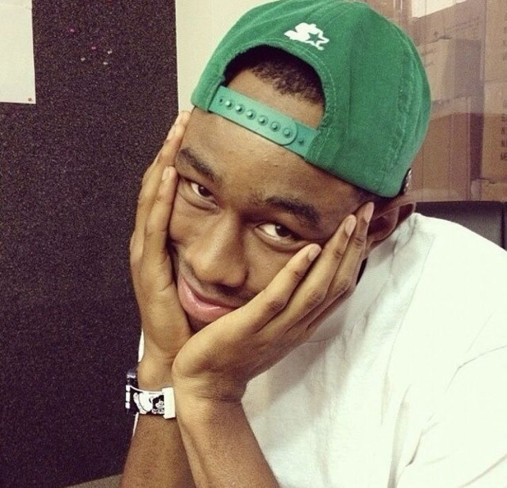 a young man wearing a green hat and holding his hands to his face while looking at the camera