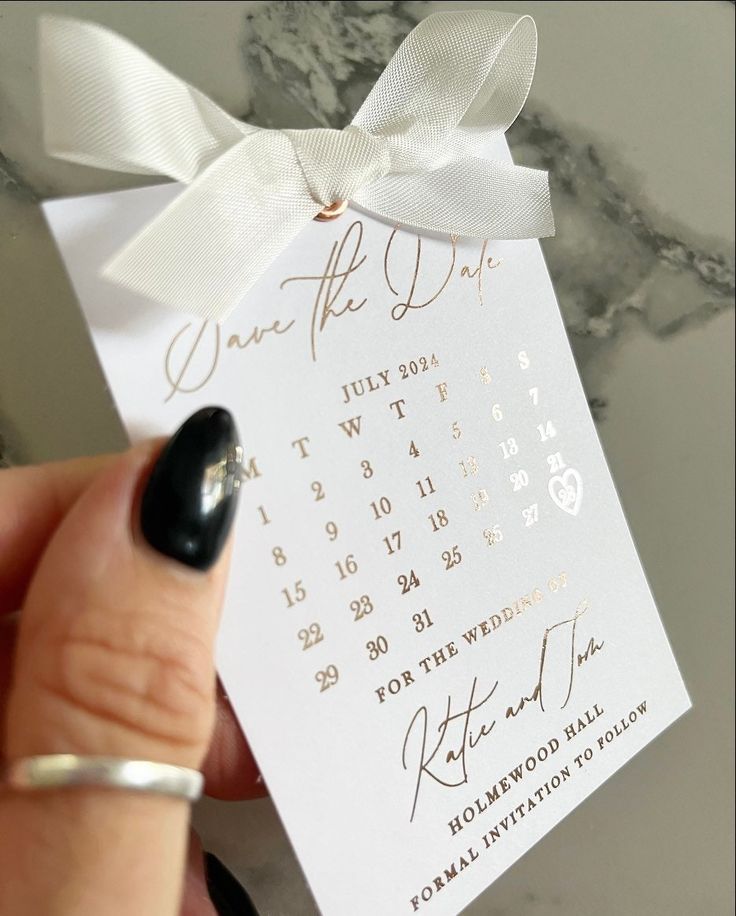 a hand holding a white and gold save the date card with a bow on it