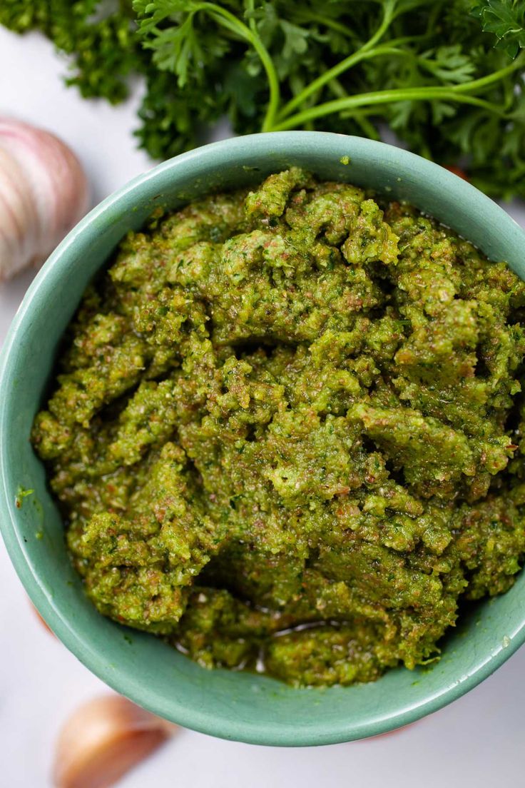 a blue bowl filled with green pesto next to garlic and parsley on a white surface