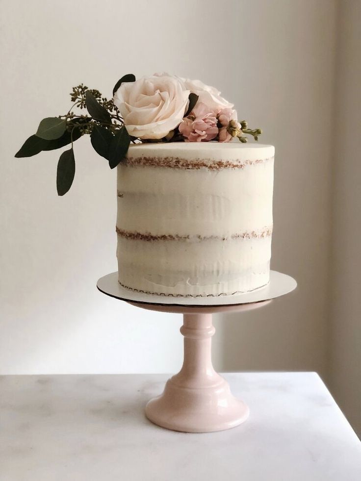 a white cake topped with flowers and greenery on a pedestal next to a wall