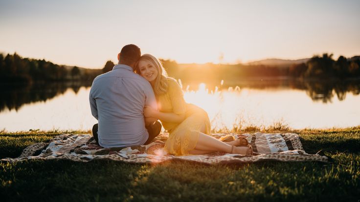 a man and woman sitting on top of a blanket next to a lake at sunset