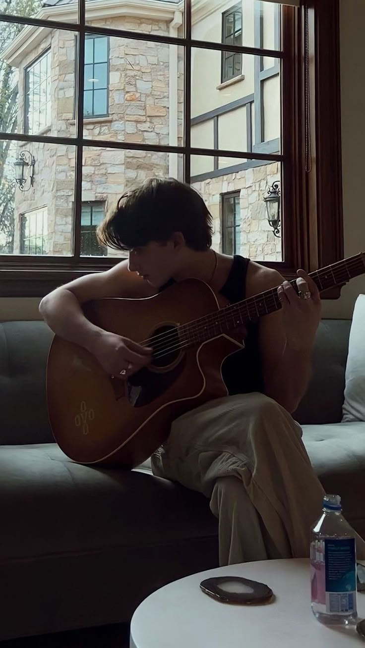 a person sitting on a couch with a guitar in front of a window and a bottle of water