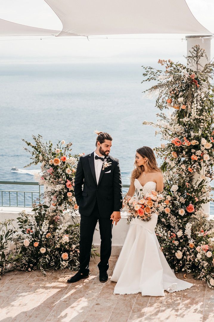 a bride and groom holding hands under an arch with flowers on it, overlooking the ocean