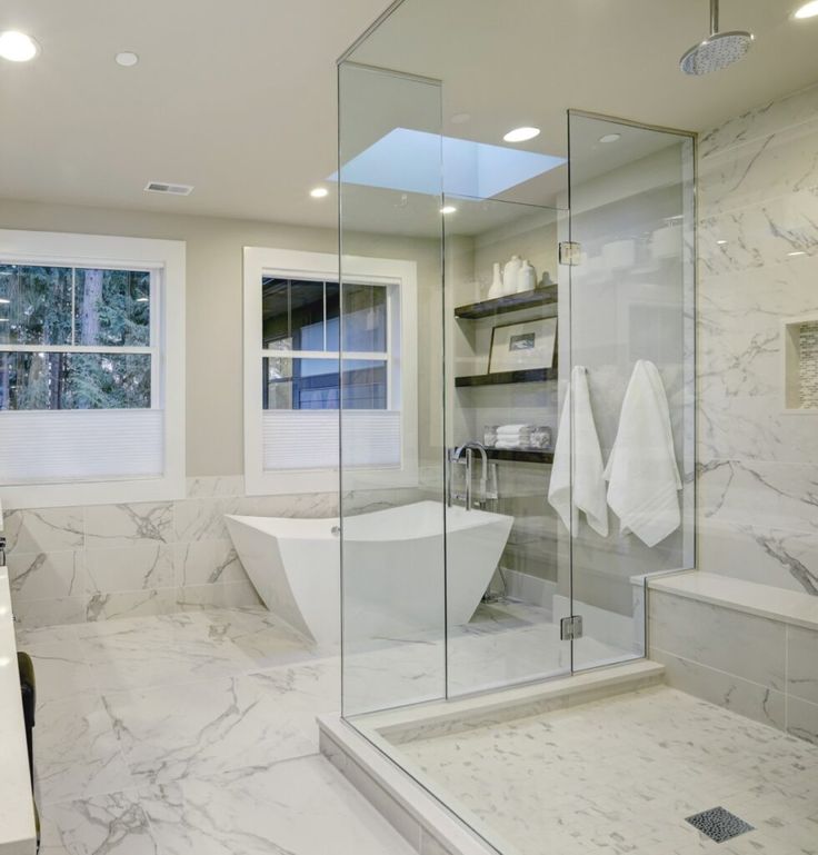 a bathroom with marble walls and flooring has a large glass shower stall, white bathtub, and sink