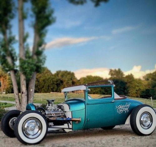 an old model car is parked on the side of the road in front of some trees