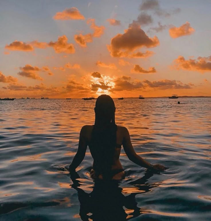 a woman is sitting in the water with her back turned to the camera as the sun sets