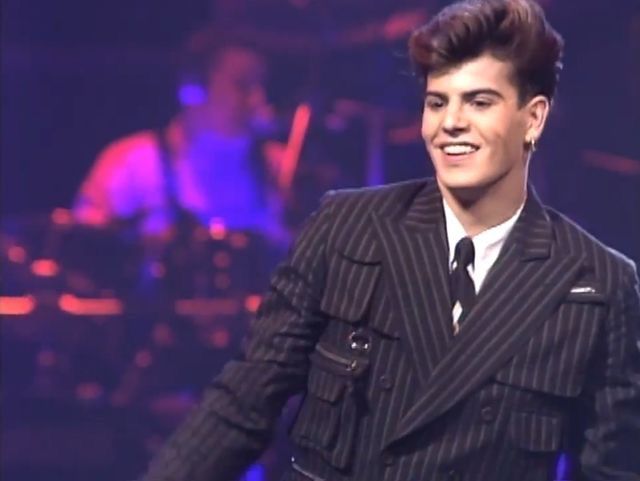 a man in a suit and tie on stage with his hands in his pockets, smiling at the camera