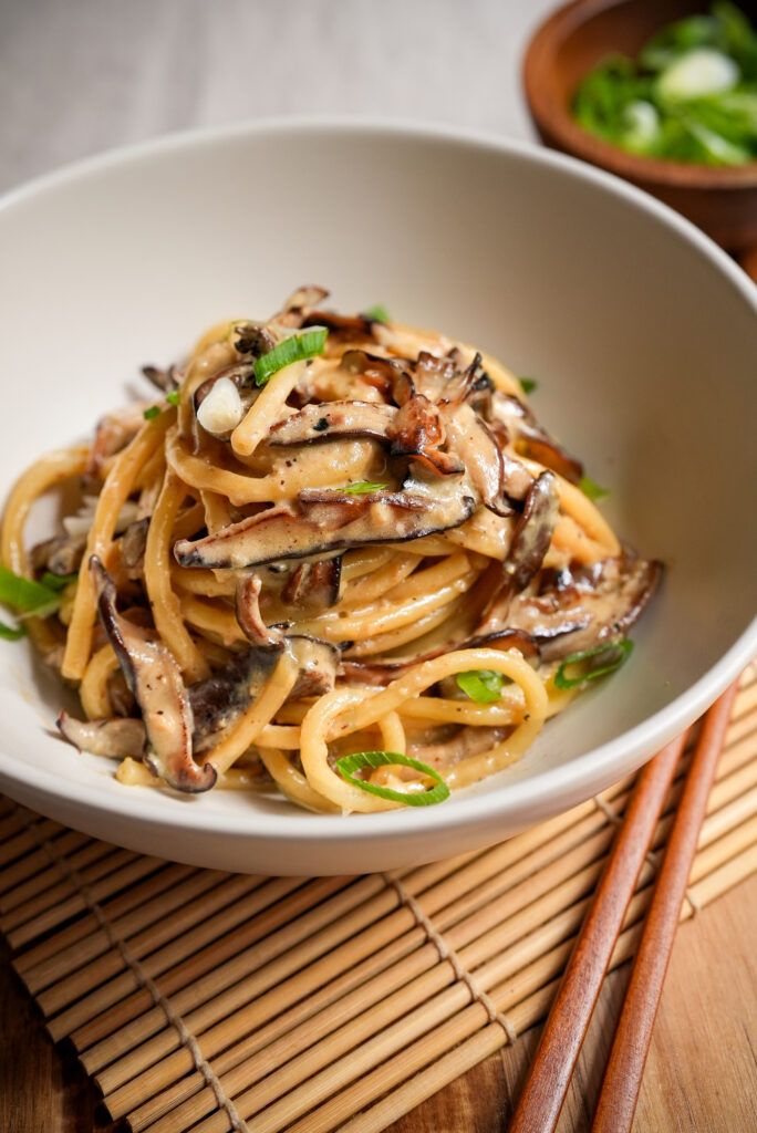 a white bowl filled with pasta and mushrooms on top of a table next to chopsticks