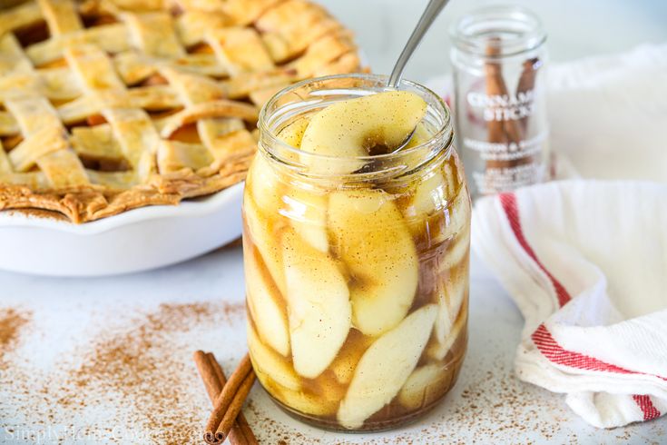 an apple pie in a glass jar with cinnamon sticks and two other desserts behind it