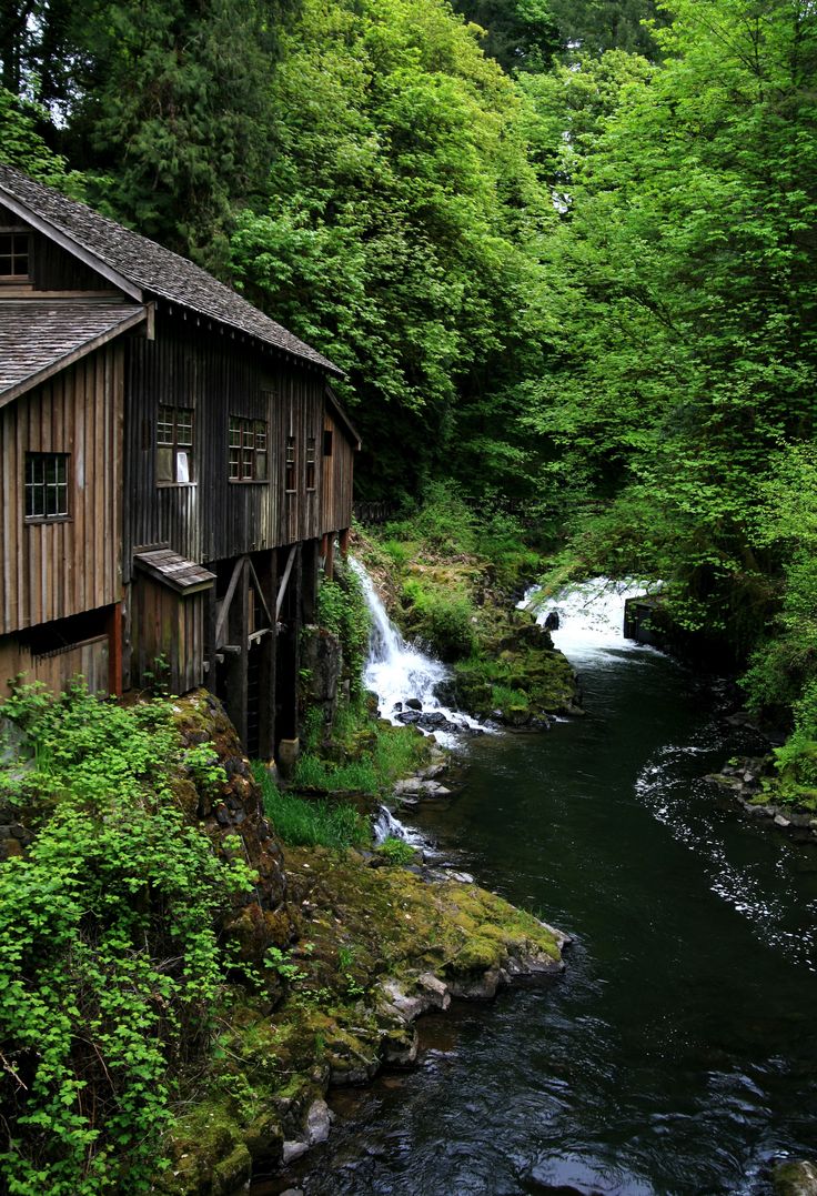 a wooden building sitting next to a river surrounded by trees and water flowing down it's side