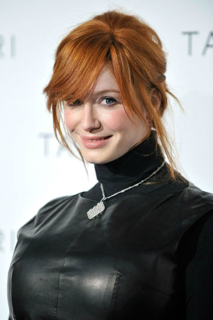 a woman with red hair wearing a black leather dress and smiling at the camera while standing in front of a white wall