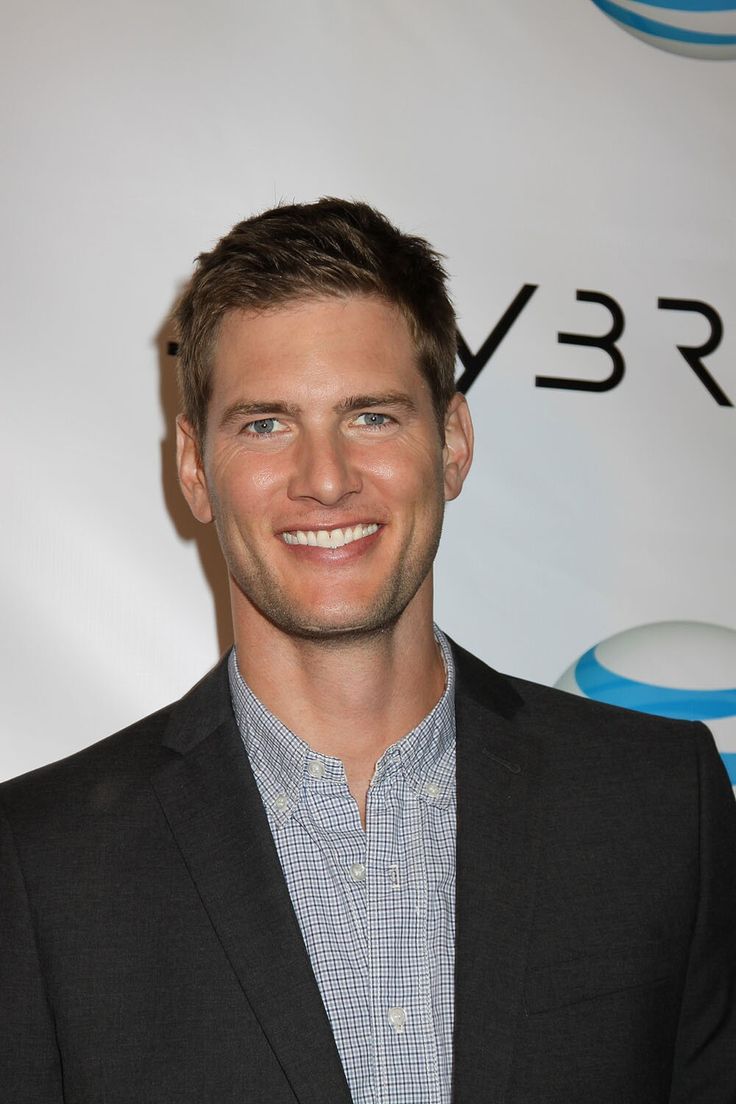 a smiling man in a suit and plaid shirt at an event with his name on the wall behind him