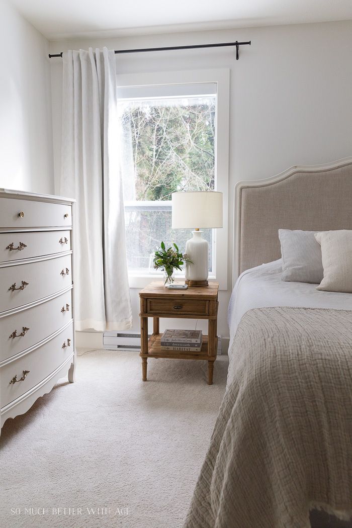 a bedroom with a bed, dresser and window in the corner that has white curtains