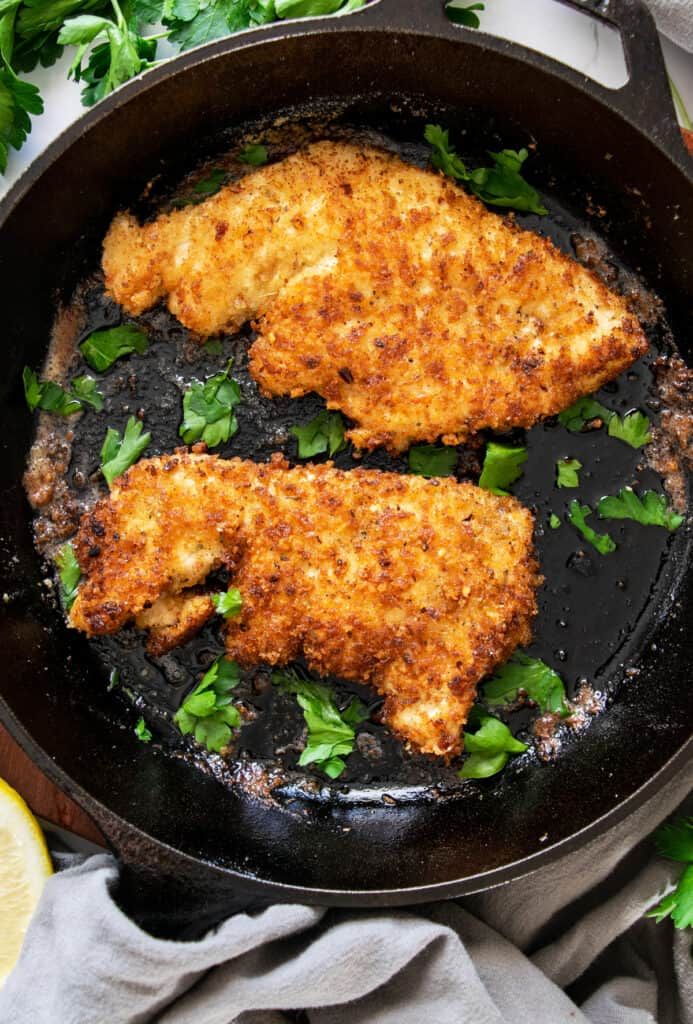 two pieces of chicken in a skillet with parsley on the side and lemon wedges next to it