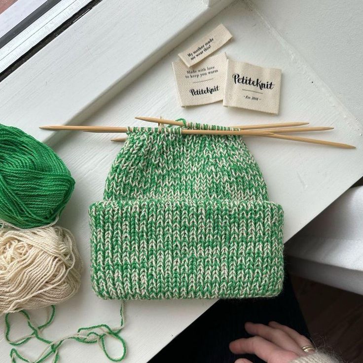 green and white knitted beanie next to yarn with knitting needles on the table