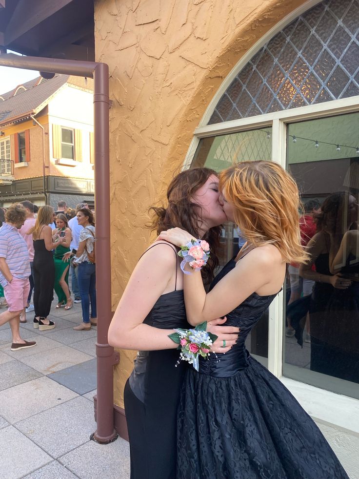 two women are kissing each other in front of a building with people standing around it