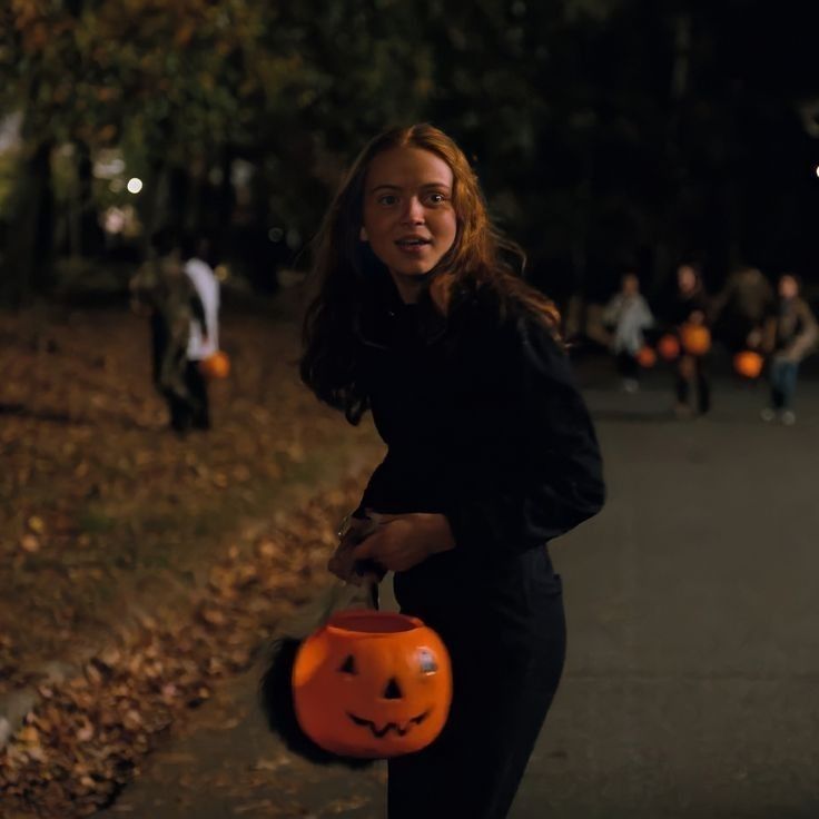 a woman holding a pumpkin bucket in the street with people walking behind her at night