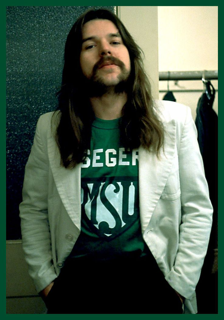 a man with long hair wearing a green shirt and white blazer standing in front of a closet