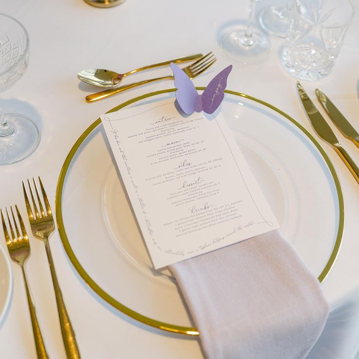 a place setting with silverware and purple butterflies on the napkins, gold rimmed cutlery