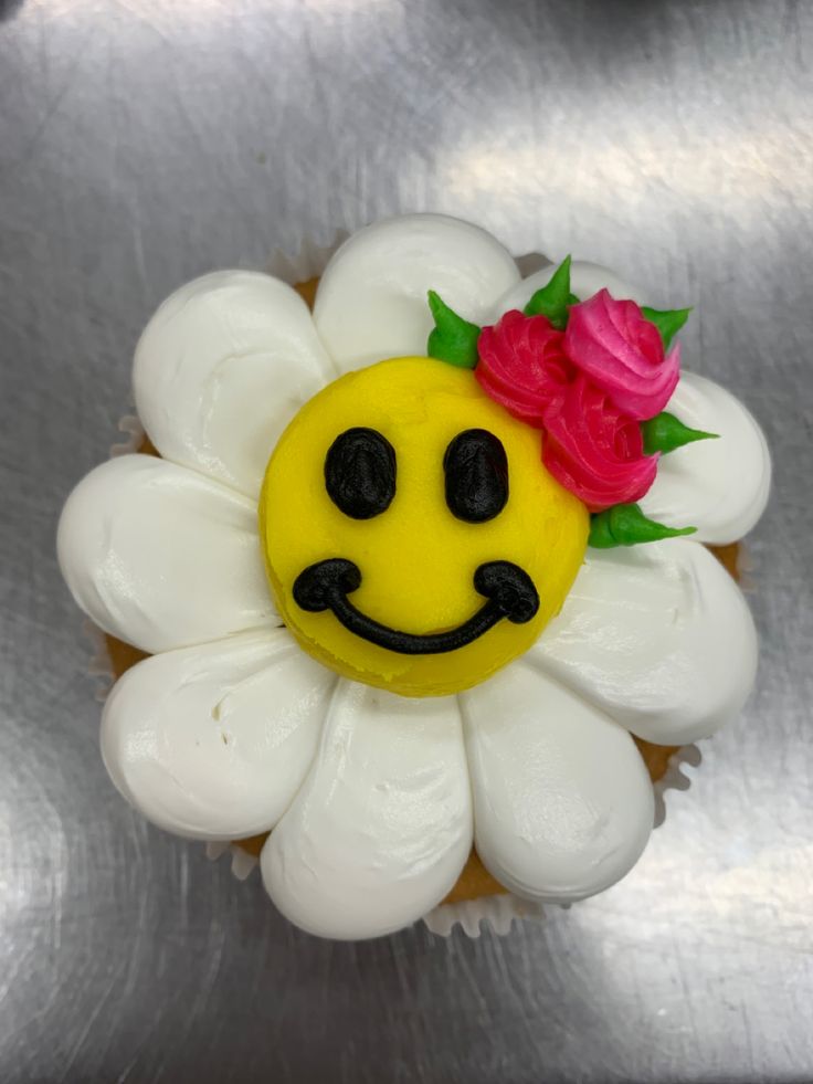 a cupcake with a smiley face and flower on it's top, sitting on a metal surface