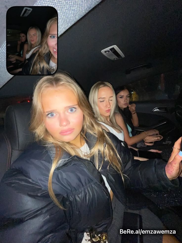 four women sitting in the back seat of a car and one is taking a selfie with her cell phone