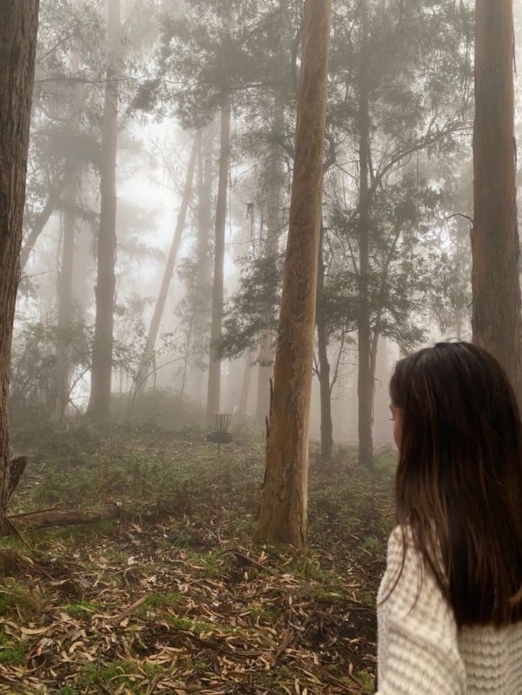 a woman standing in the middle of a forest on a foggy day with trees