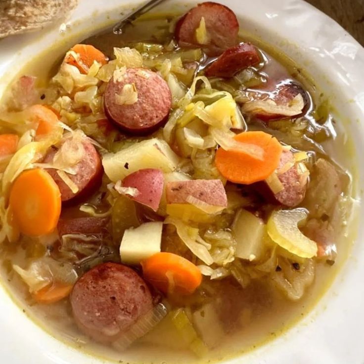 a white bowl filled with cabbage and sausage soup next to a piece of bread on top of a wooden table