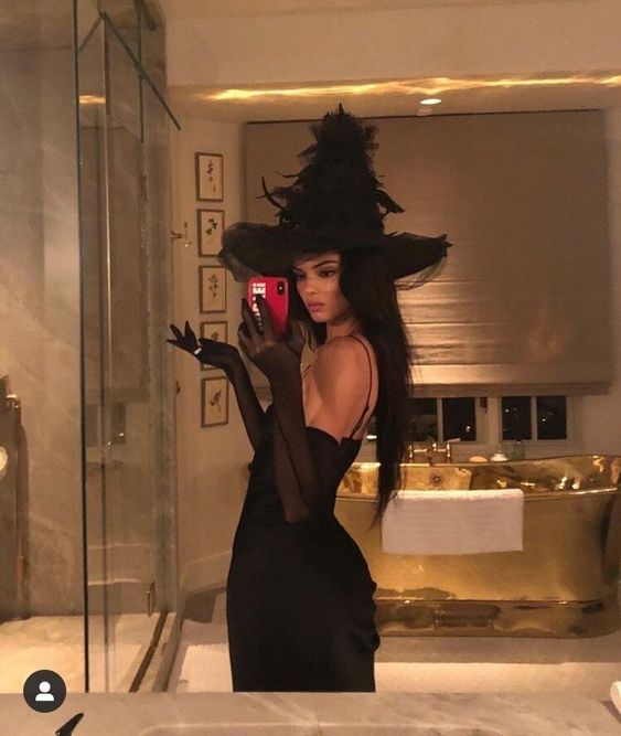 a woman in a black dress and hat taking a selfie with her cell phone