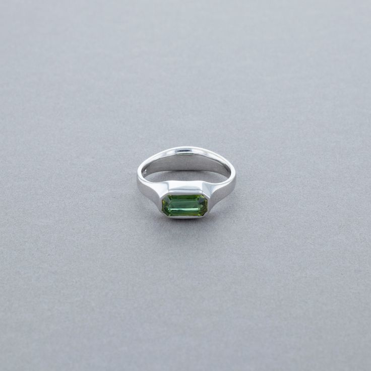 This lovely sapphire ring can be worn as a statement piece, a stack-able ring, or as a non-traditional engagement ring! The sapphire is dark green in color, and emerald cut. It is bezel set, and lies horizontally in a 14K White Gold band. The stone is about 4.5MM from top to bottom, and approximately 8MM from side to side. The band tapers with a 3MM width beside the stone, and a thicker width at the bottom of the ring. We are certain you will love and appreciate your ring, but if you are not com Modern Green Emerald-cut Jewelry, Modern Emerald Rings With Baguette Cut, Modern Baguette Cut Emerald Promise Ring, Green Sapphire Baguette Cut Ring, Modern Baguette Cut Emerald Ring, Silver And Green Engagement Ring, Modern Sterling Silver Emerald Cut Sapphire Ring, Classic Green Ring With Smooth Bezel, Modern Bezel-set Emerald Ring For Anniversary