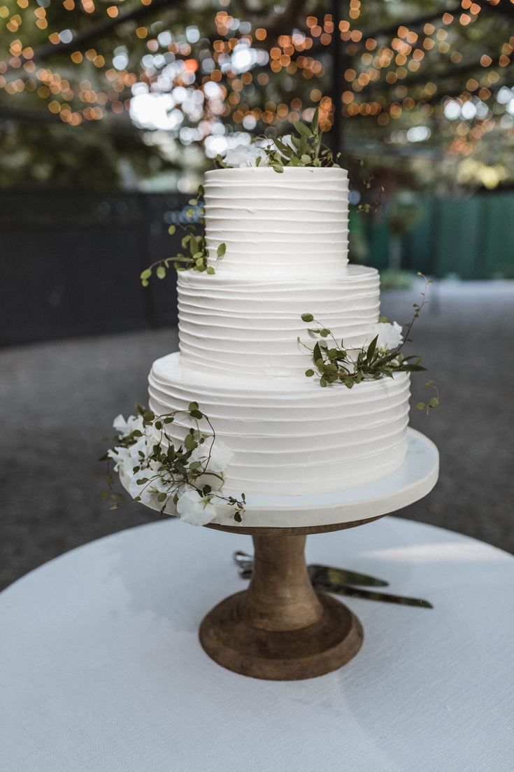 a white wedding cake sitting on top of a round table with greenery and lights in the background