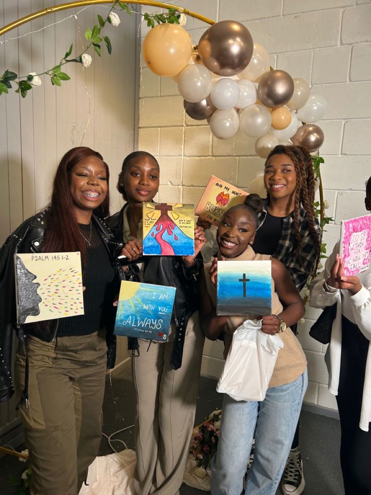 four women holding up paintings in front of a white brick wall with balloons and streamers