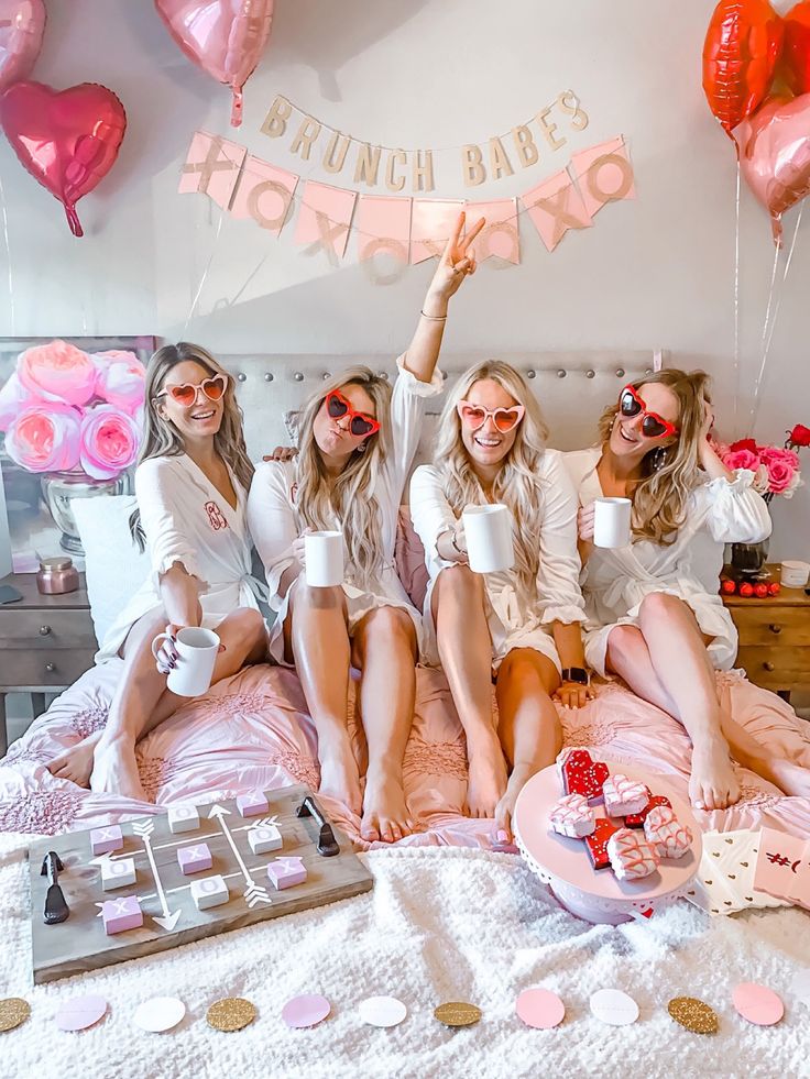 three beautiful women sitting on top of a bed with pink decorations and balloons hanging from the ceiling