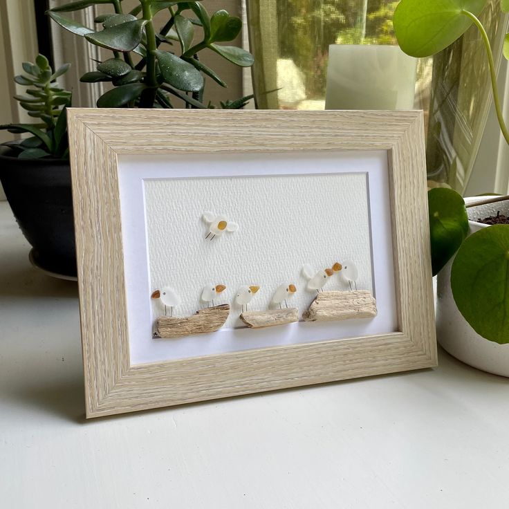 three little white sheep are standing in front of a framed photo on a table next to a potted plant