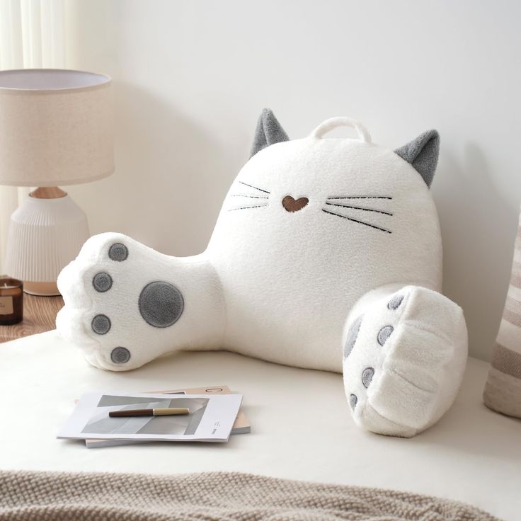 a white cat pillow sitting on top of a bed next to a book and lamp