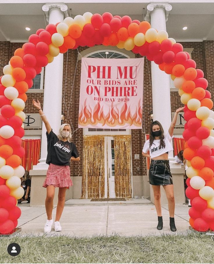 two women standing in front of a building with an orange and white arch decorated with balloons