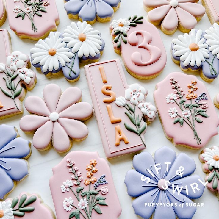 decorated cookies with flowers and letters are displayed on a white tablecloth covered in pink, blue, and yellow icing