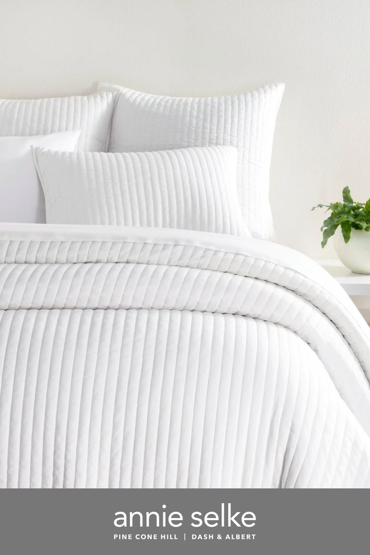 a bed with white comforter and pillows on it next to a potted plant