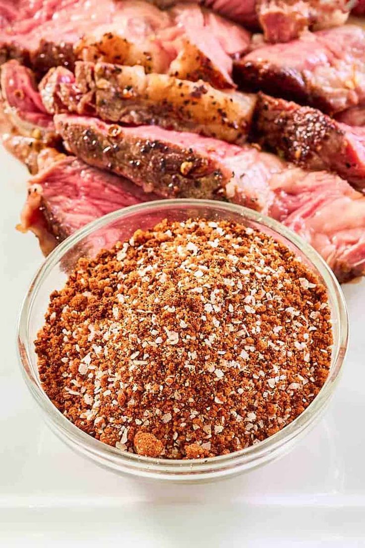 steak seasoning in a glass bowl next to sliced meat on a white counter top