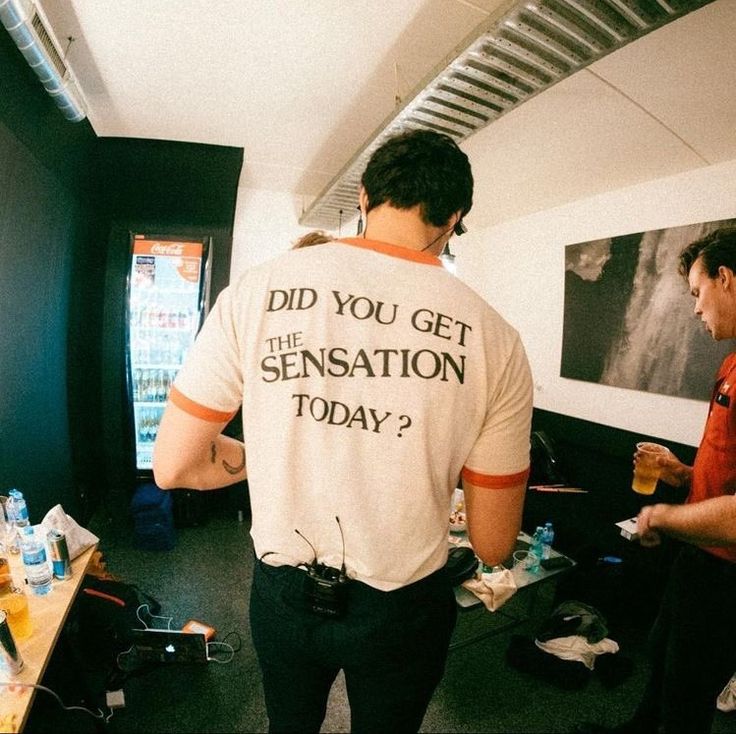 two men standing in an office talking to each other while one man is wearing a t - shirt that says did you get the sensation today?