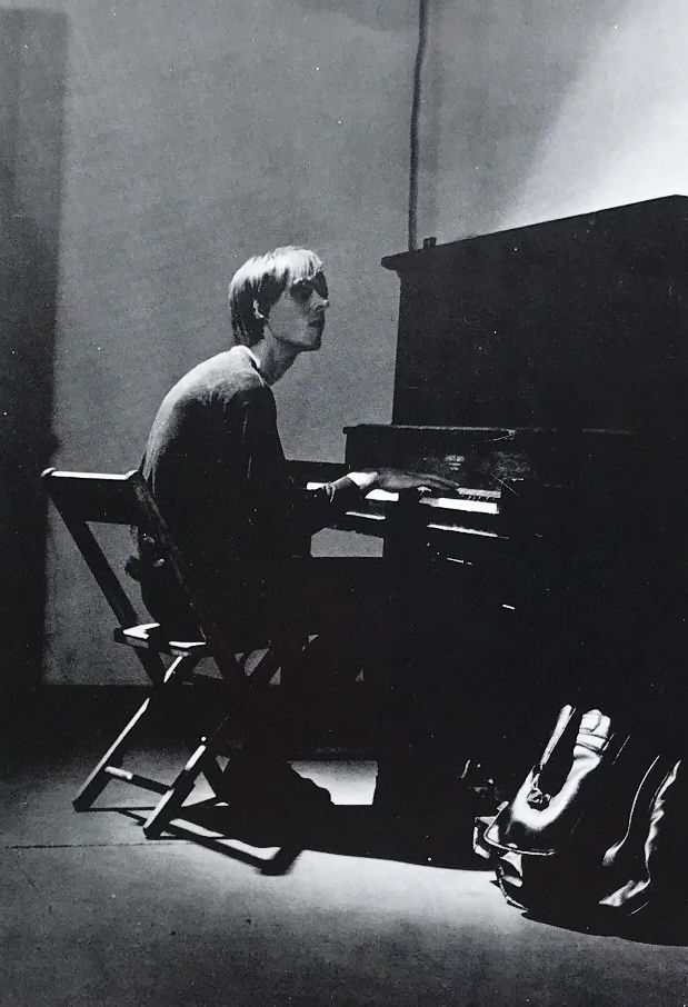 a young man sitting at a piano in front of a black and white photo with shoes on the floor