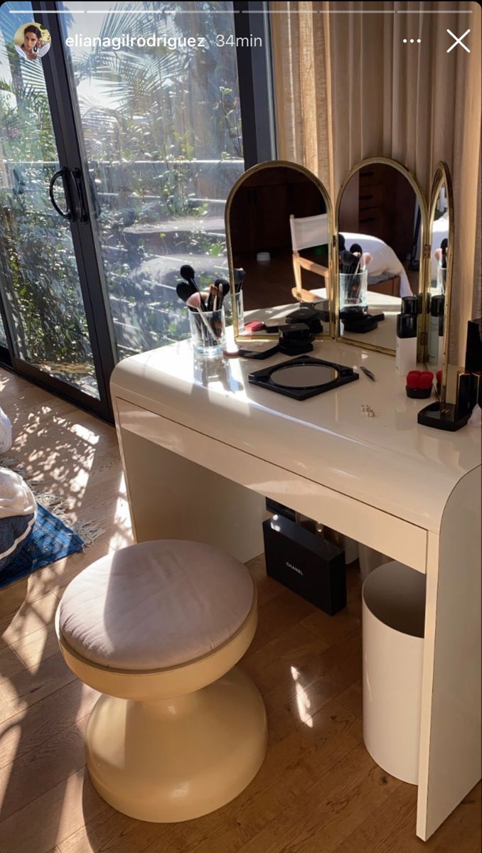 a white vanity table with two mirrors and a stool in front of it, next to a large window